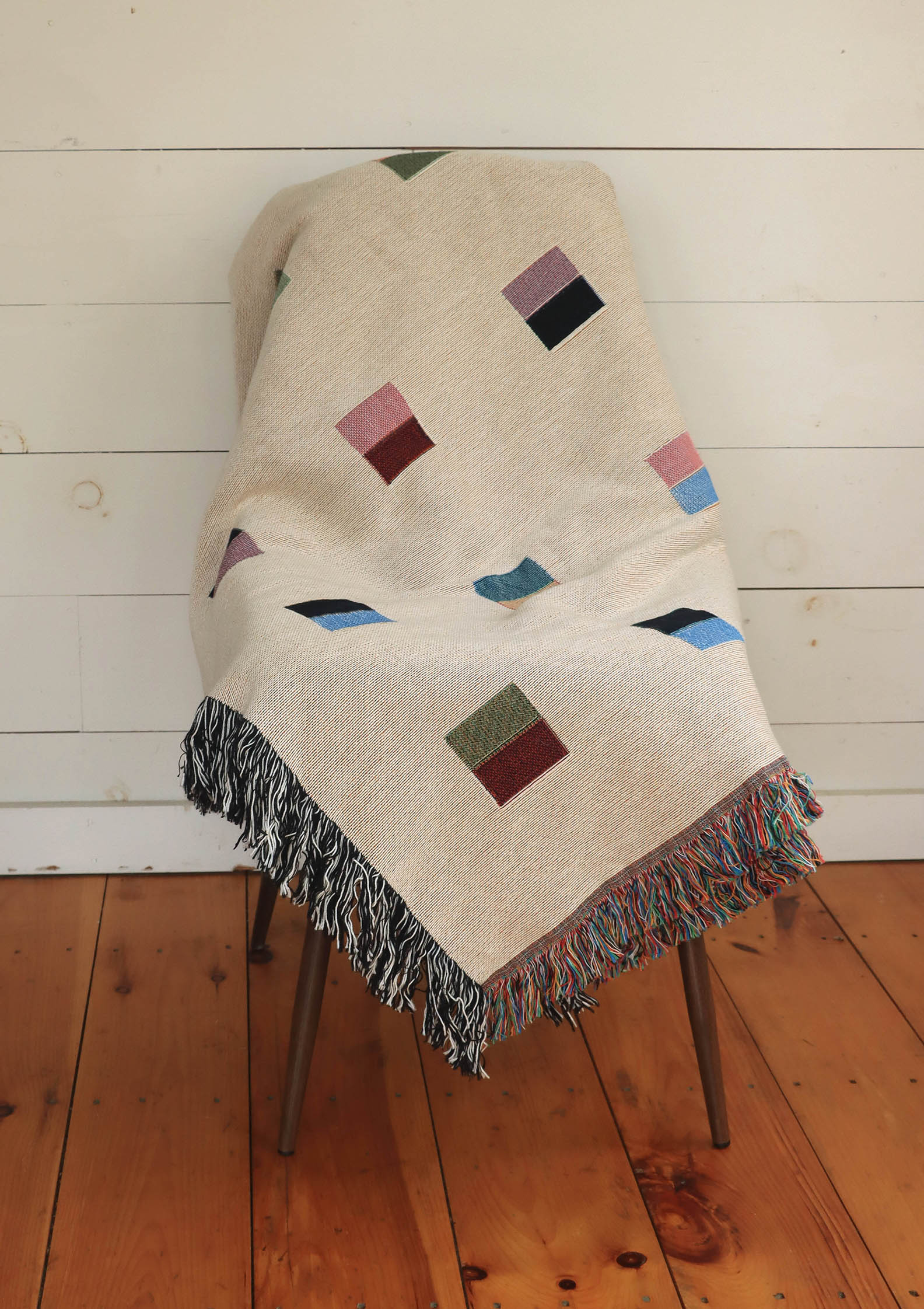 A cream-colored woven blanket with a Modern geometric split squares pattern of small multicolored squares and fringe edges. Scandinavia Design style for contemporary home decor.