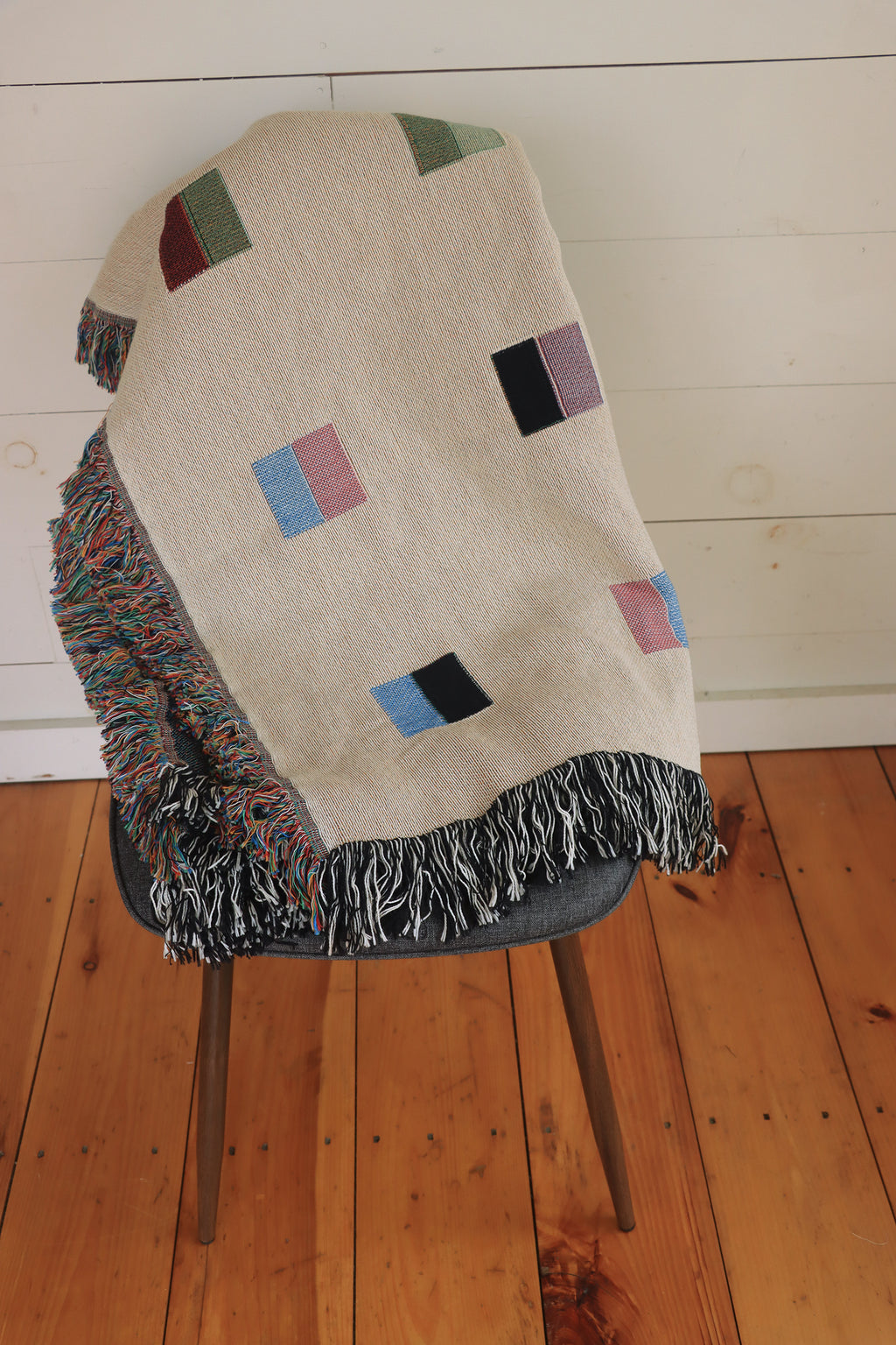 A cream-colored woven blanket with a Modern geometric split squares pattern of small multicolored squares and fringe edges. Scandinavia Design style for contemporary home decor.