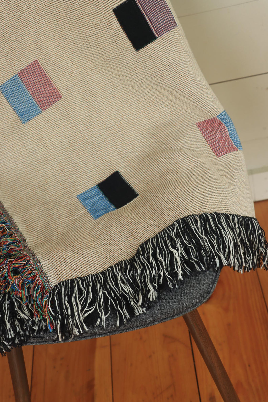 A cream-colored woven blanket with a Modern geometric split squares pattern of small multicolored squares and fringe edges. Scandinavia Design style for contemporary home decor.