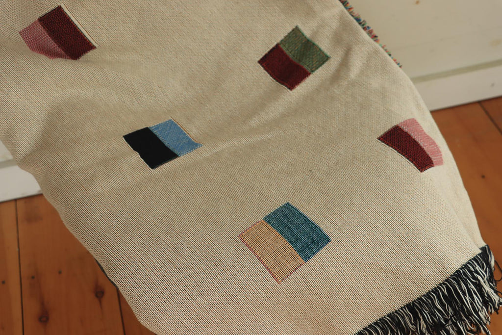 A cream-colored woven blanket with a Modern geometric split squares pattern of small multicolored squares and fringe edges. Scandinavia Design style for contemporary home decor.