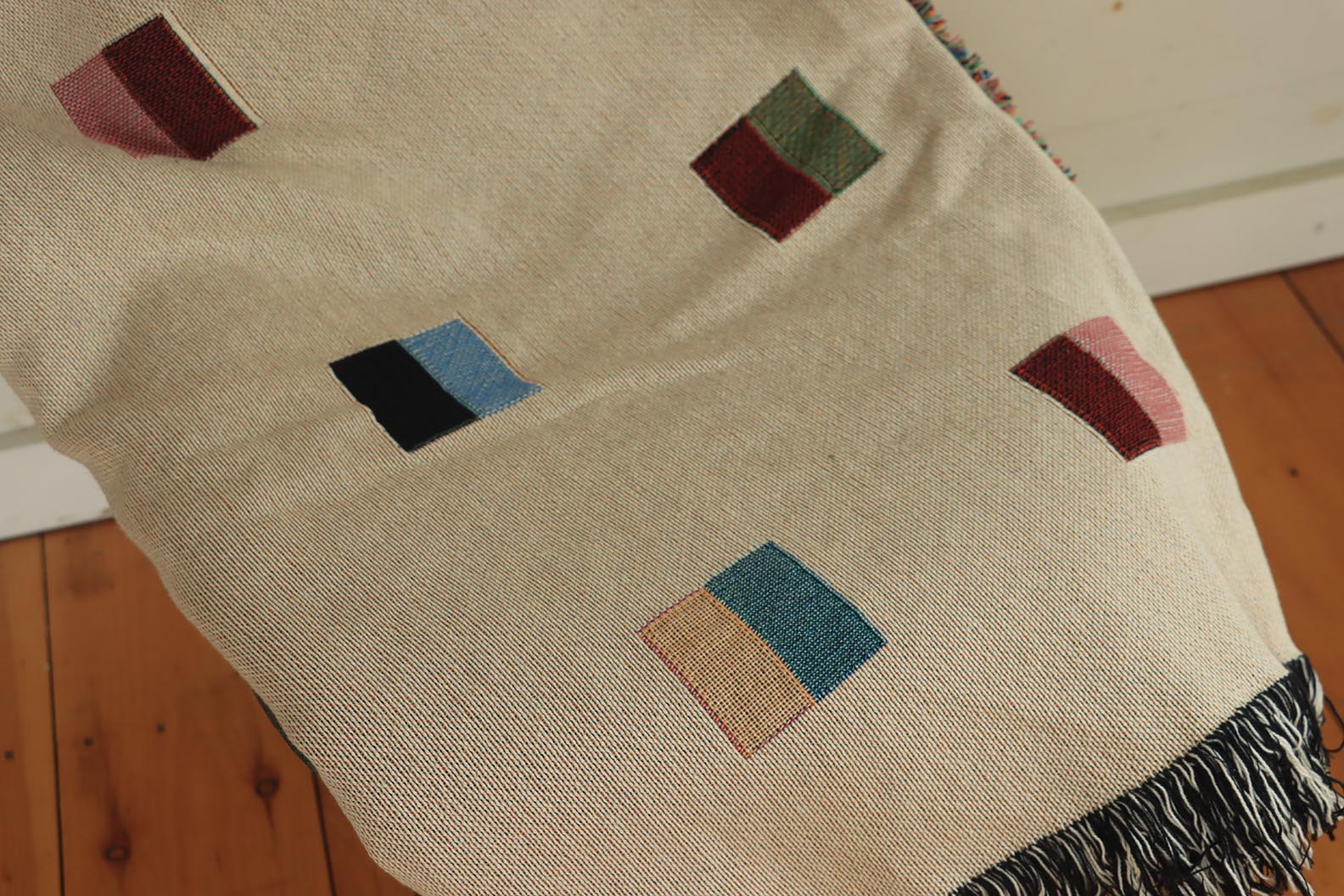 A cream-colored woven blanket with a Modern geometric split squares pattern of small multicolored squares and fringe edges. Scandinavia Design style for contemporary home decor.