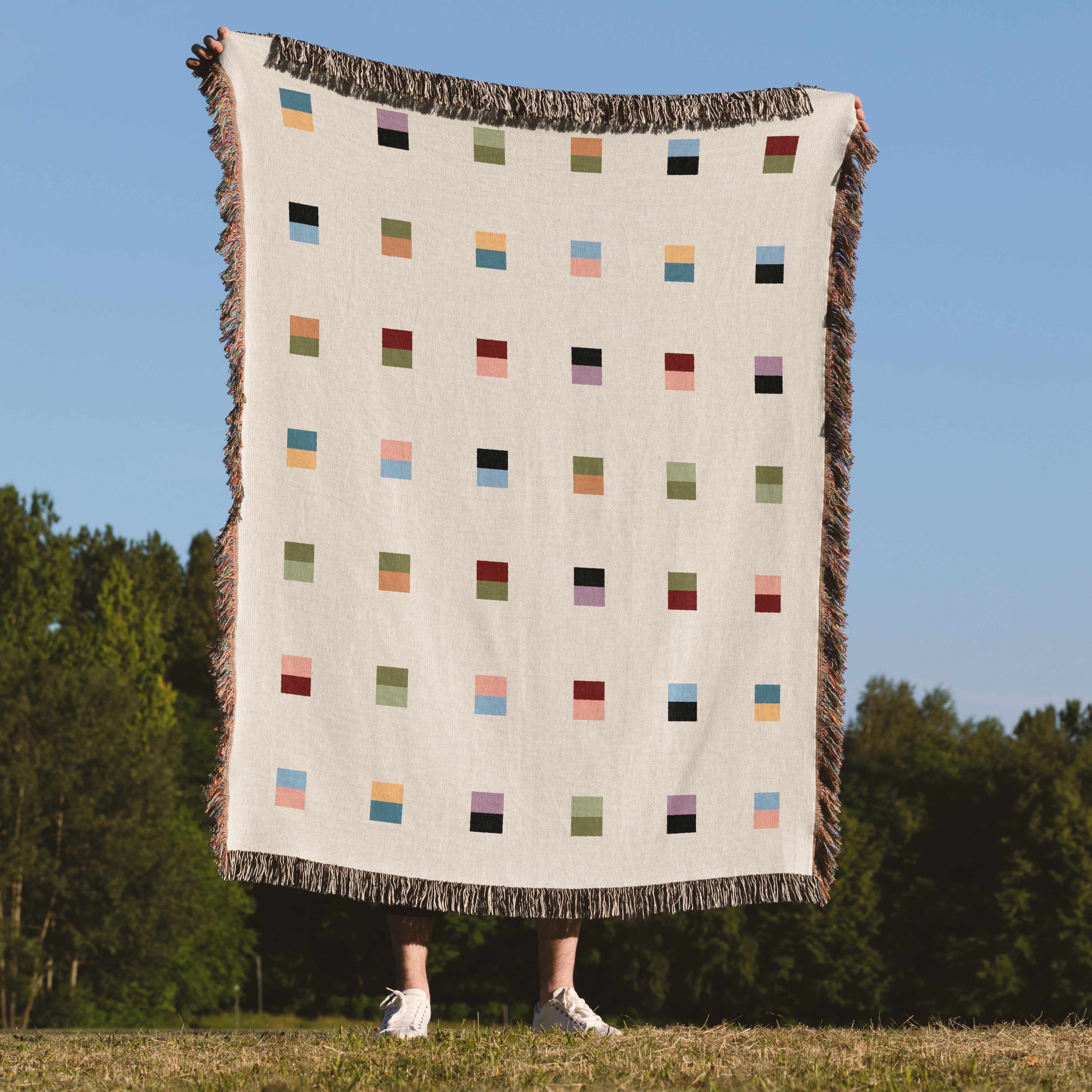 A cream-colored woven blanket with a Modern geometric split squares pattern of small multicolored squares and fringe edges. Scandinavian Design style for contemporary home decor.