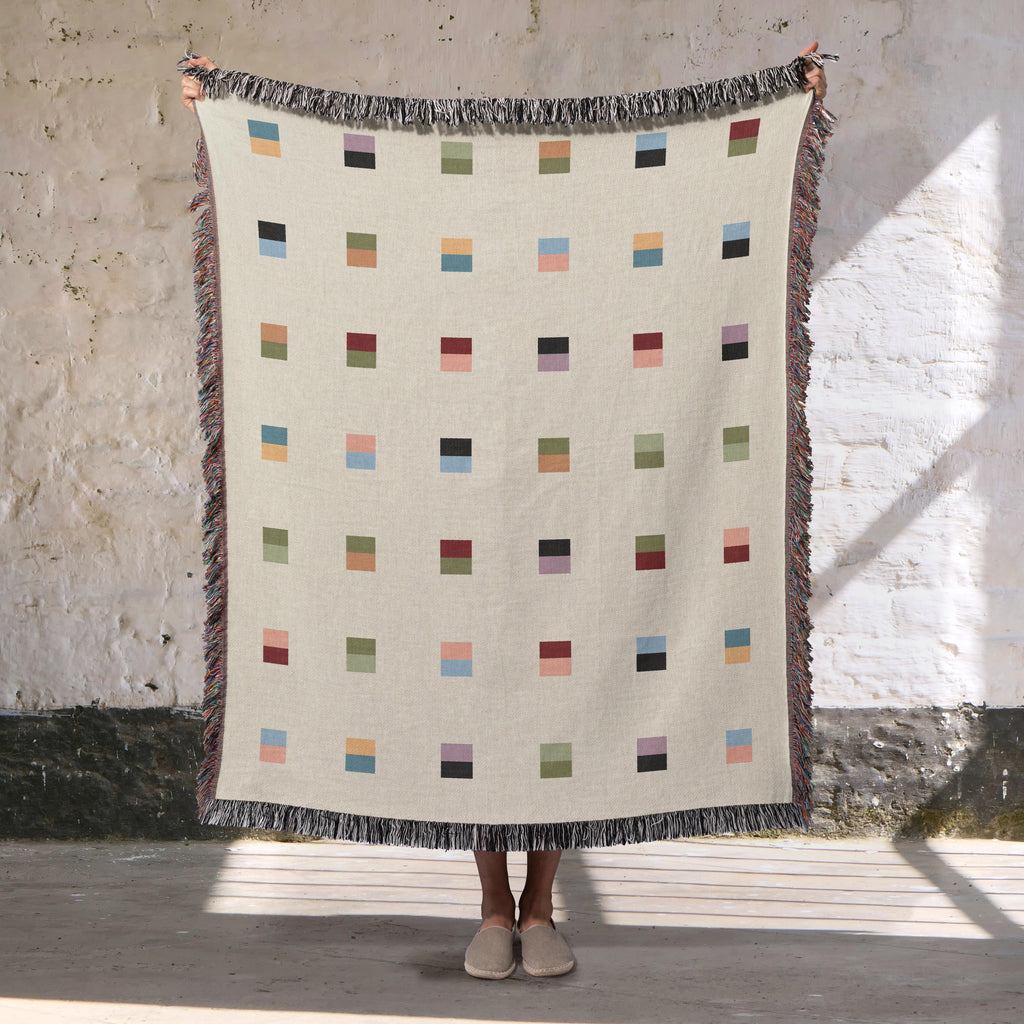 A cream-colored woven blanket with a Modern geometric split squares pattern of small multicolored squares and fringe edges. Scandinavia Design style for contemporary home decor.