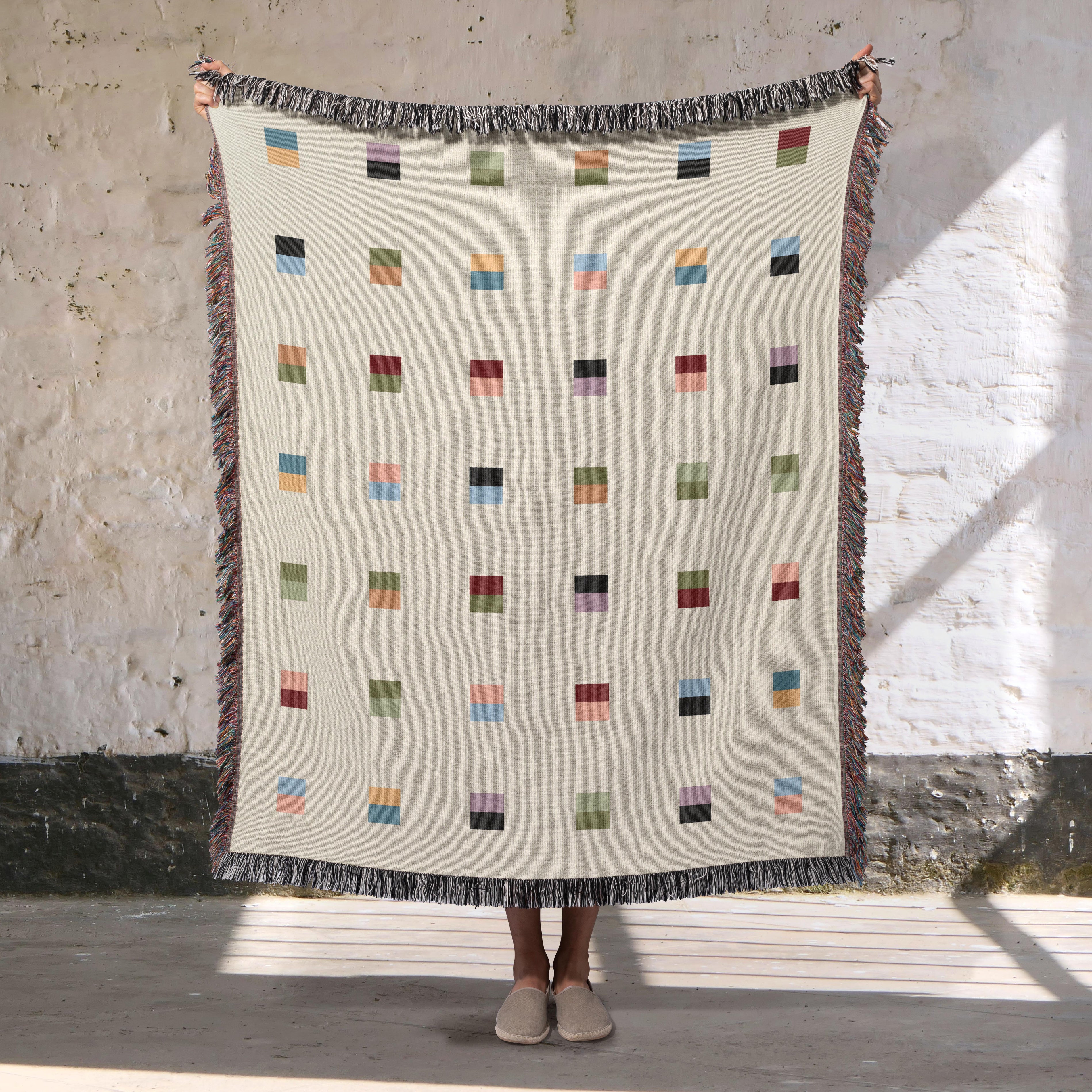 A cream-colored woven blanket with a Modern geometric split squares pattern of small multicolored squares and fringe edges. Scandinavia Design style for contemporary home decor.