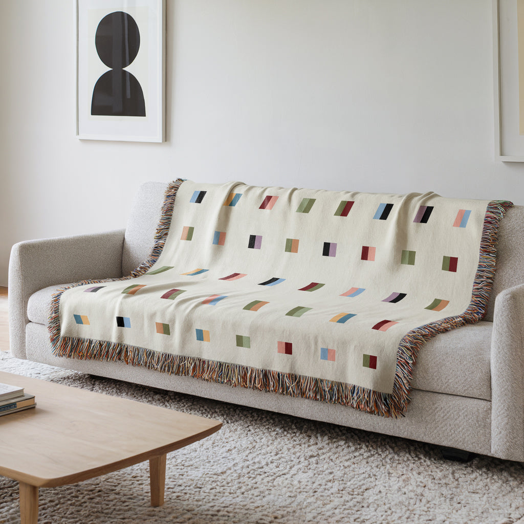 A cream-colored woven blanket with a Modern geometric split squares pattern of small multicolored squares and fringe edges. Scandinavia Design style for contemporary home decor.